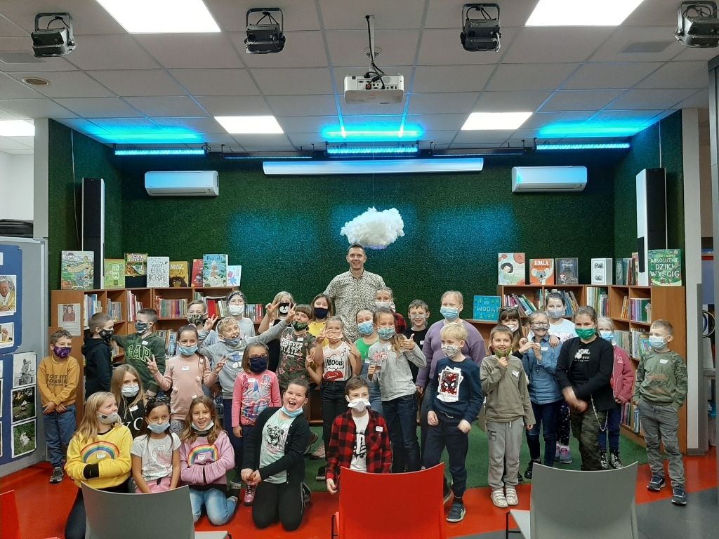 Autor książek dla dzieci Rafał Witek oraz grupa trzydziesty dzieci stoją na tle zielonej ściany, a nad nimi wisi biała chmurka