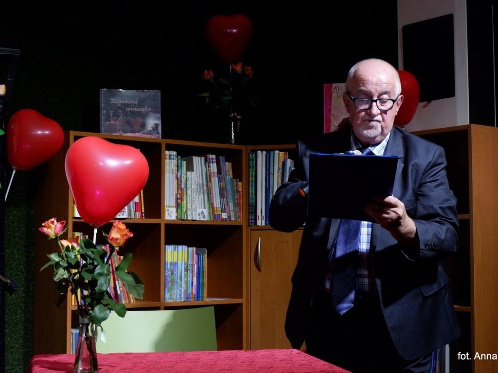 Mężczyzna w okularach czytający tekst. Po jego lewej stronie widać dwa czerwone balony w kształcie serca. W tel biblioteczka z książkami.