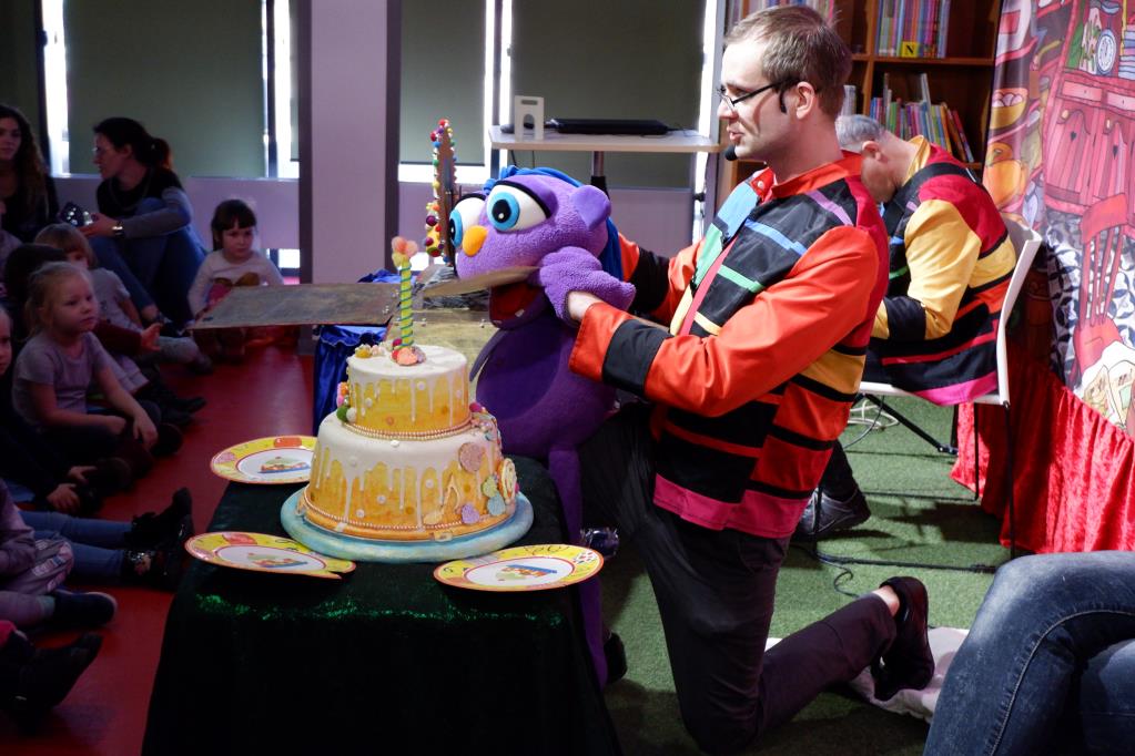 Aktor z założoną na rękę maskotką Muzolka klęczy przy stoliku, na którym znajduje się duży tort urodzinowy