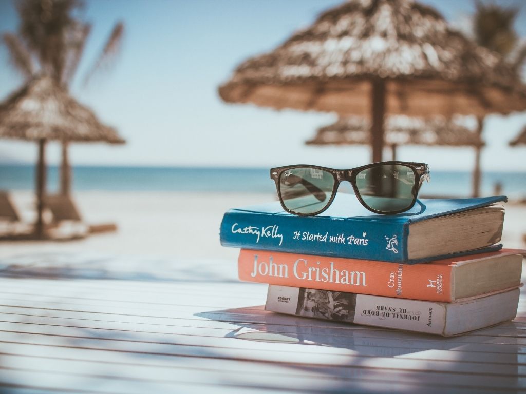 Okulary przeciwsłoneczne położone na stosie książek. W tle parasole na plaży.