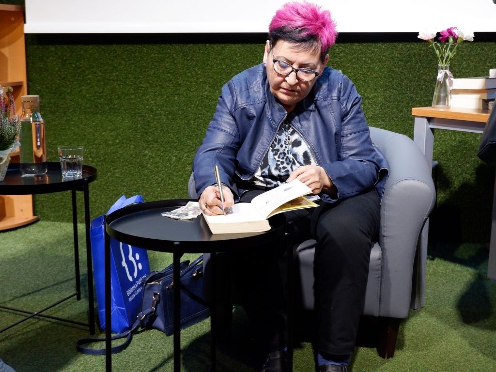 Kobieta w czarno-różowych włosach siedzi w szarym fotelu i podpisuje książkę.