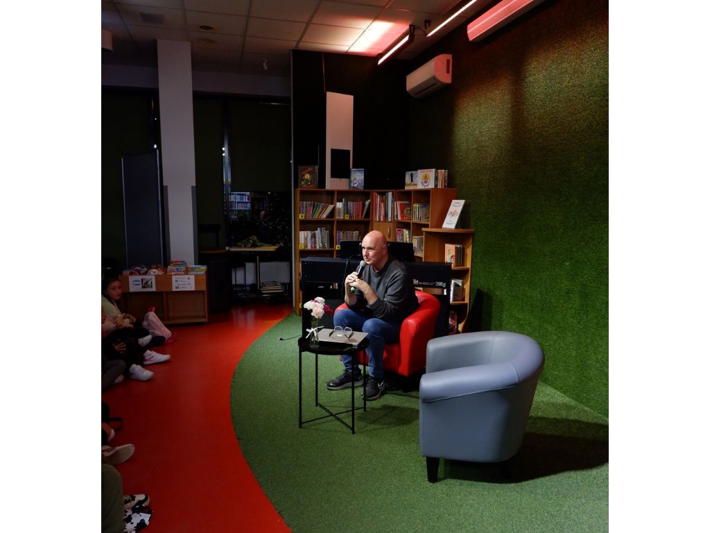 Mężczyzna w ciemnym swetrze siedzi na czerwonym fotelu, w ręku trzyma mikrofon. W tle regały z książkami.